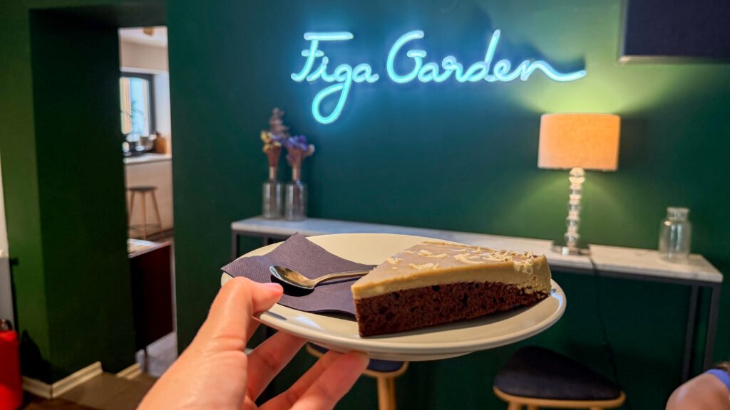 Figa Garden by Henrik Zagreb | Krista the Explorer Slice of cake on a plate held by a person in a cafe, with a blue neon sign in front.