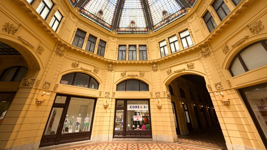 Ornate yellow indoor shopping arcade with arched storefronts and a glass skylight.