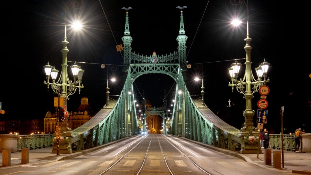 Night view of a green ornate suspension bridge with tall decorative towers and bright street lamps.