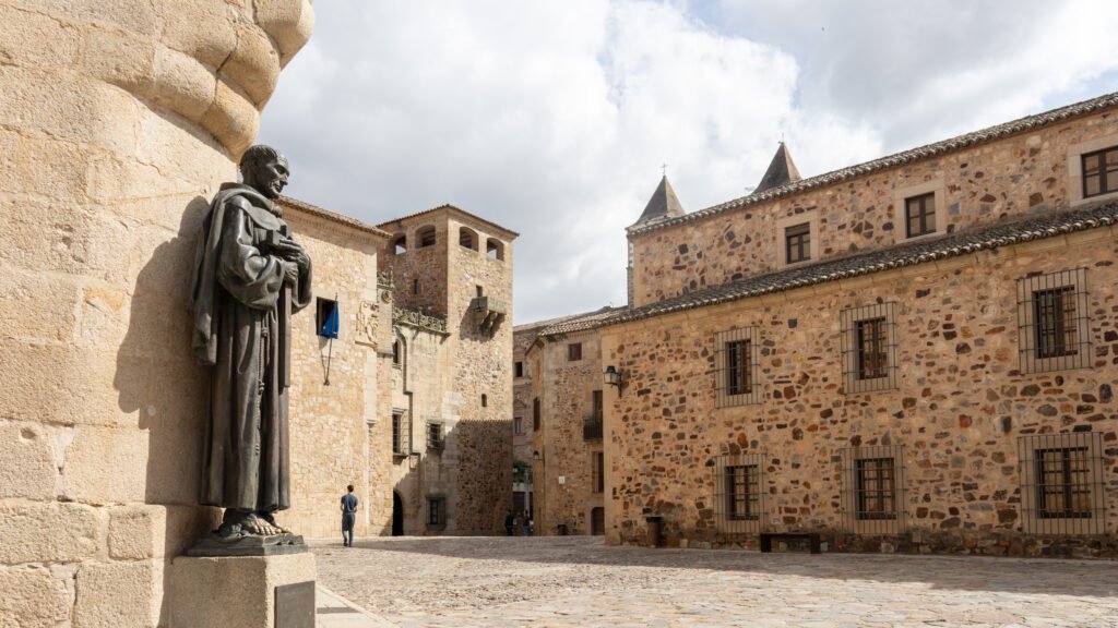Plaza de Santa María Cáceres | Krista the Explorer Statue in a sunlit medieval plaza, with stone buildings and a cobblestone surface nearby.