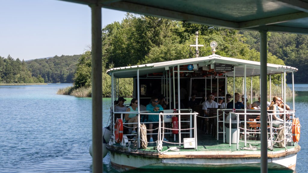 Plitvice Lakes | Krista the Explorer People ride a covered ferry on a calm lake, seated at tables.