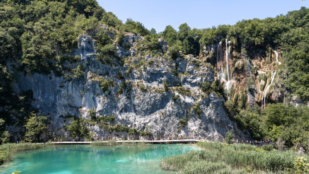 Plitvice Lakes | Krista the Explorer Turquoise lake with a wooden boardwalk along a rocky cliff and a thin waterfall.