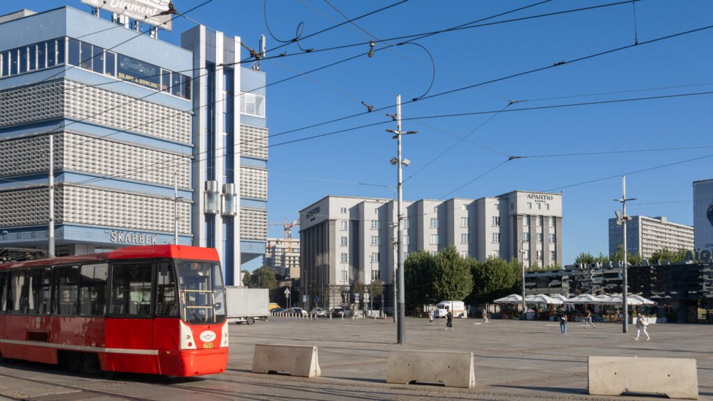 Red and white tram at a city square with blue modern buildings.