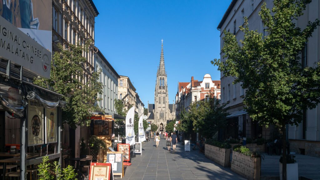 City street with outdoor cafe seating on the left, a tall Gothic church spire at the end, and trees lining the right sidewalk.