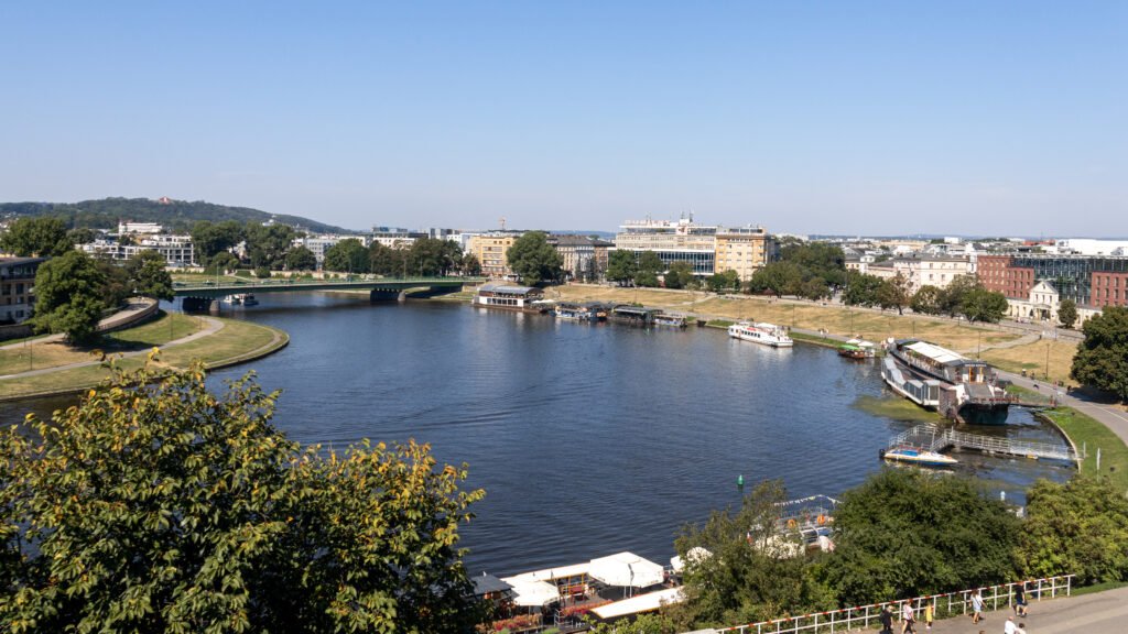 Vistula River Krakow | Krista the Explorer Urban riverside cityscape with a curved walkway, grassy park, and boats docked along the water.