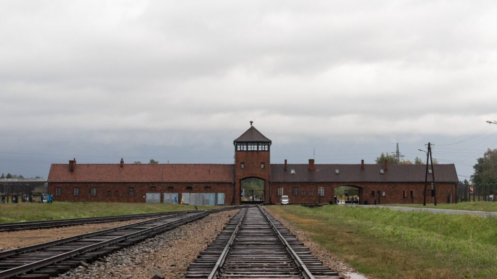 Auschwitz-Birkenau Krakow | Krista the Explorer Rail tracks converge toward a brick gatehouse with a peaked roof under a cloudy sky.