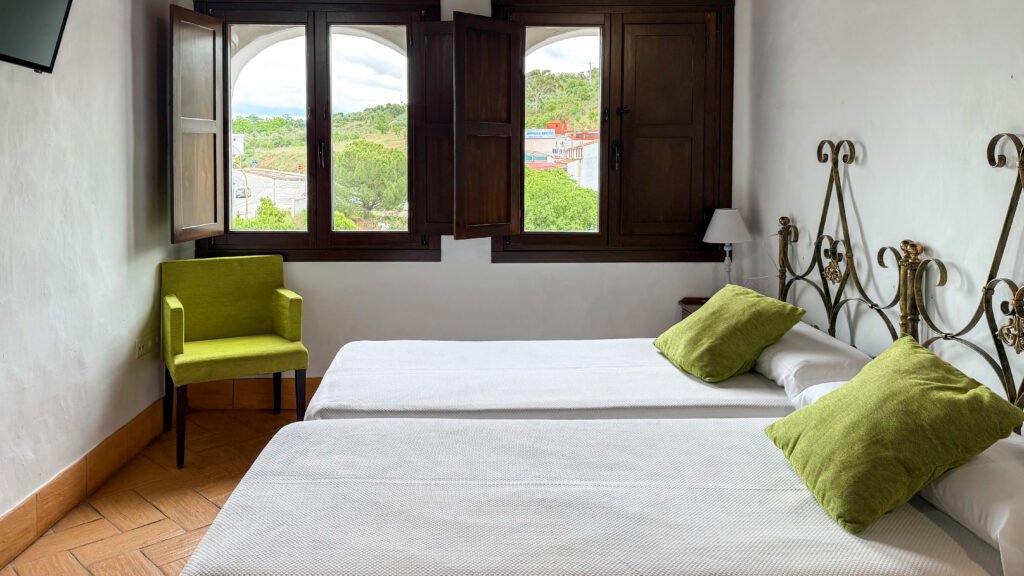 Hostal Los Alisos Caceres | Krista the Explorer Hotel room with two white beds, green pillows and a dark wood window with shutters.