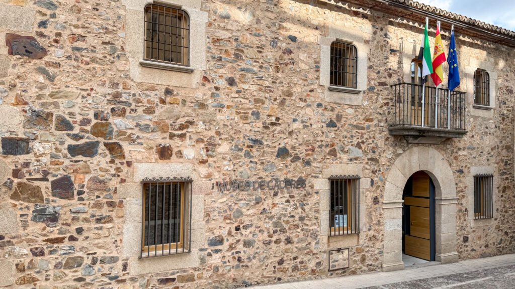 Museum of Cáceres | Krista the Explorer Historic stone building facade with barred windows and a balcony displaying three flags.