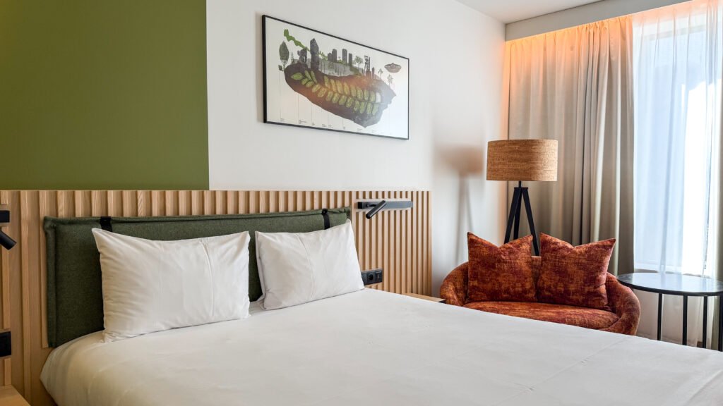 Hotel room with white bedding, a wooden slat headboard, green accent wall, and framed landscape art.