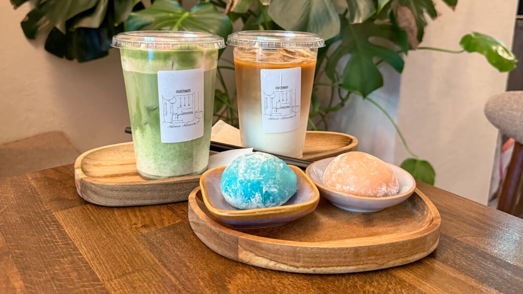 Two iced beverages in clear cups with lids on a wooden tray, plus blue and peach mochi desserts.
