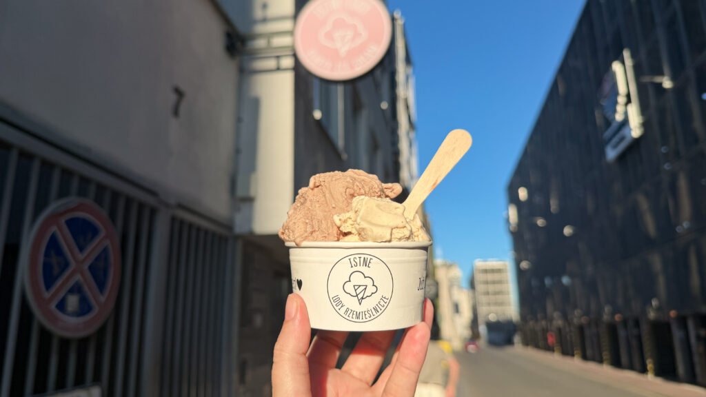 Ice cream cup with two scoops and a wooden spoon, held up against a city street.