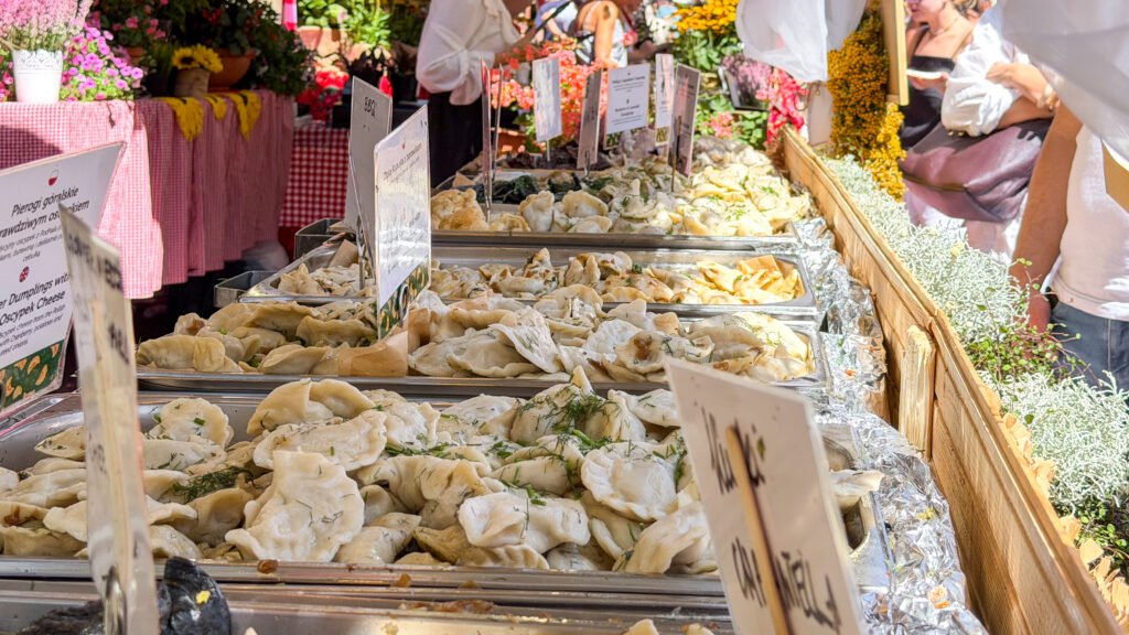 Pierogi Krakow | Krista the Explorer Market stall with trays of pierogi, signs listing flavours, and colourful flowers in the background.