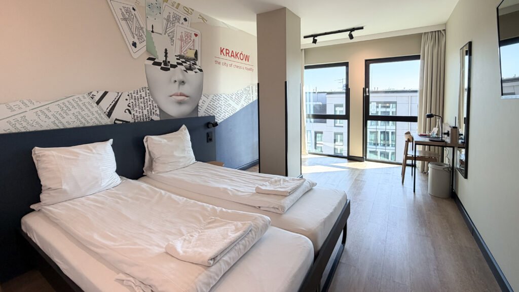 MEININGER Kraków Centrum | Krista the Explorer Hotel room with two twin beds, white linens, and a chess-themed mural including'KRAKÓW' text on the wall.