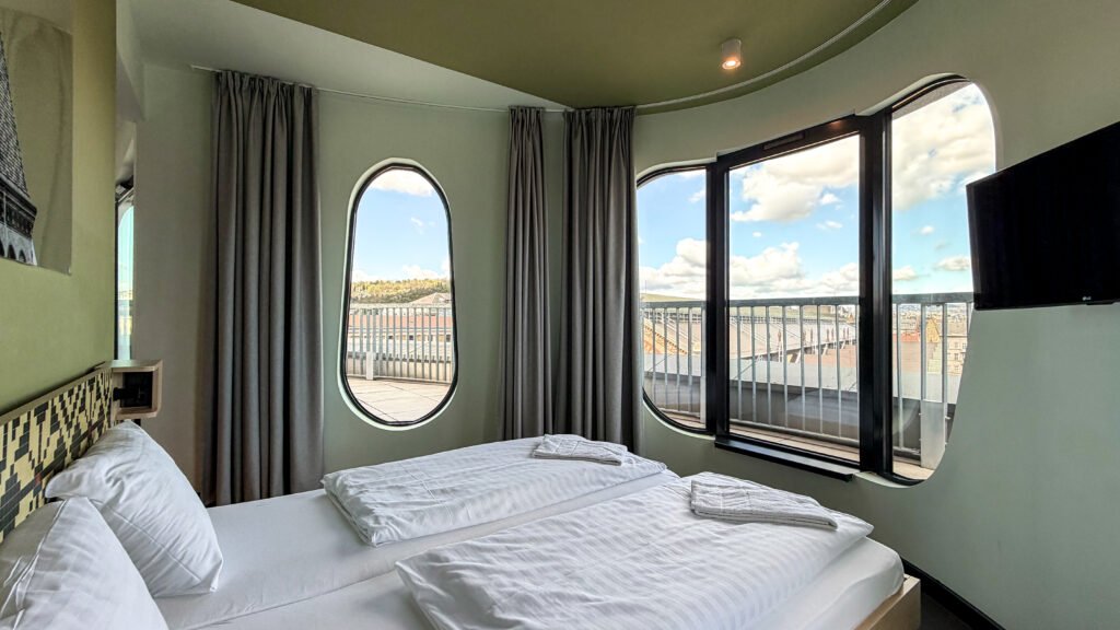 Hotel bedroom with two white beds, gray curtains, and curved wall windows overlooking a city skyline.