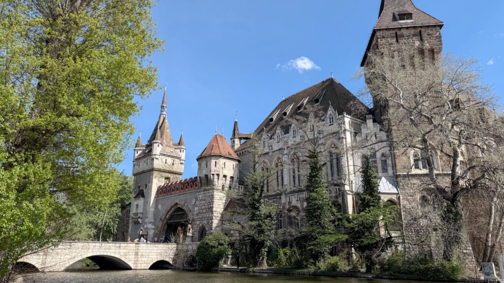 Historic stone castle with towers beside a river, arched bridge, and lush green trees.