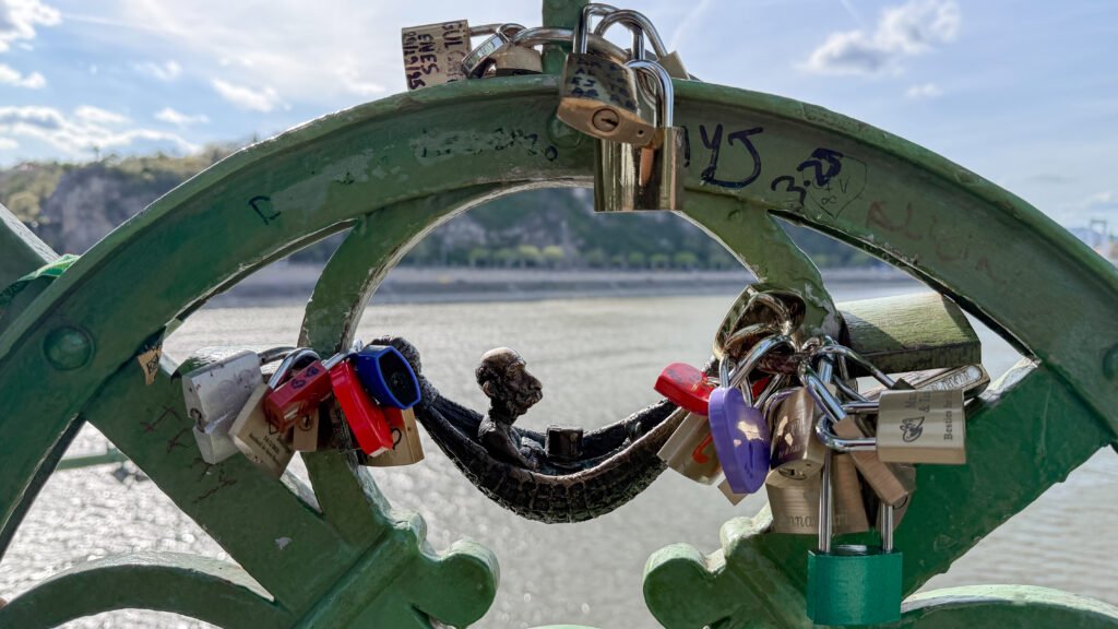 Close-up of a green railing covered in colourful padlocks and a mini statue hanging on hammock.