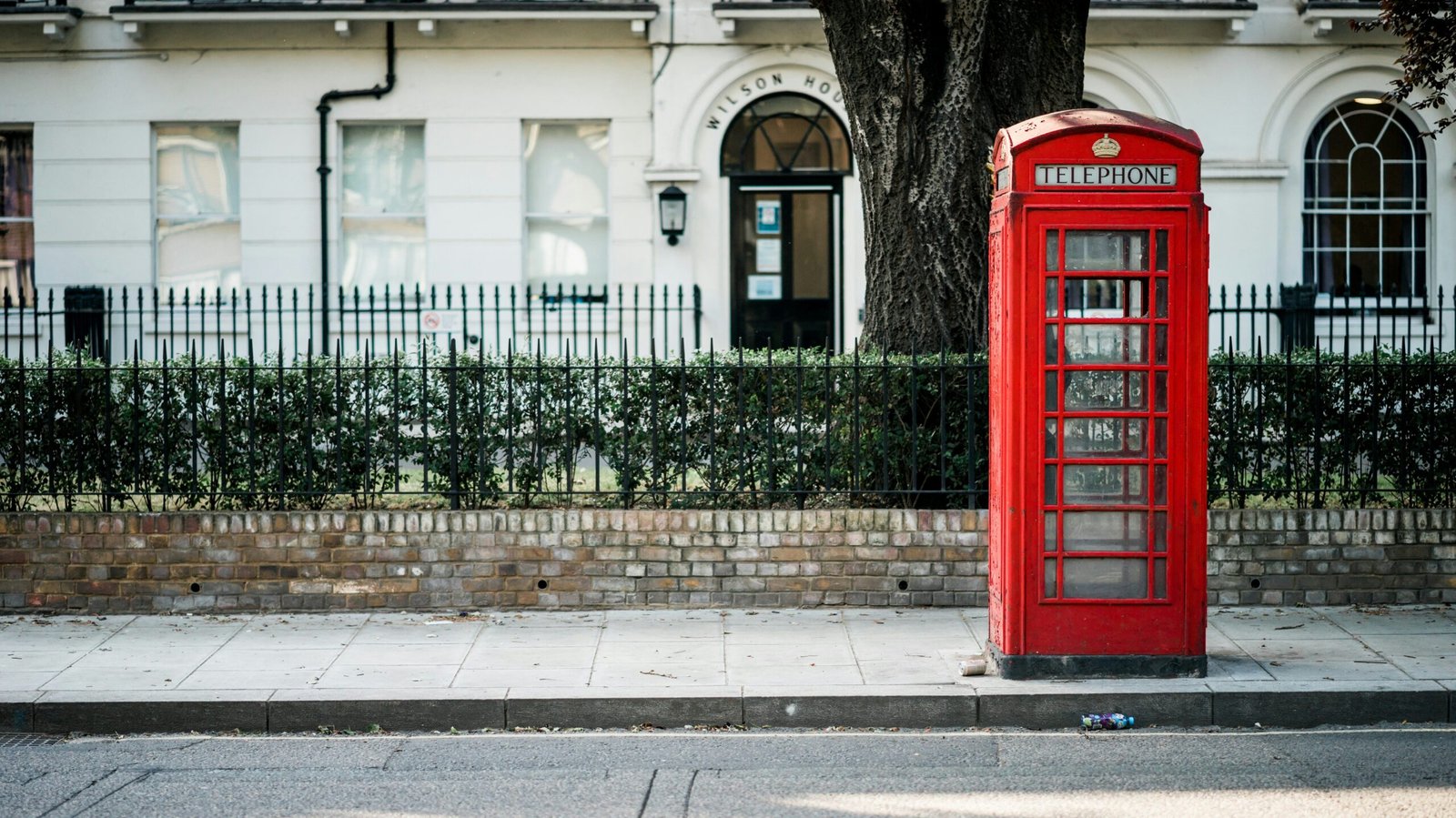 Red British-style telephone booth on a sidewalk in front of a white building.