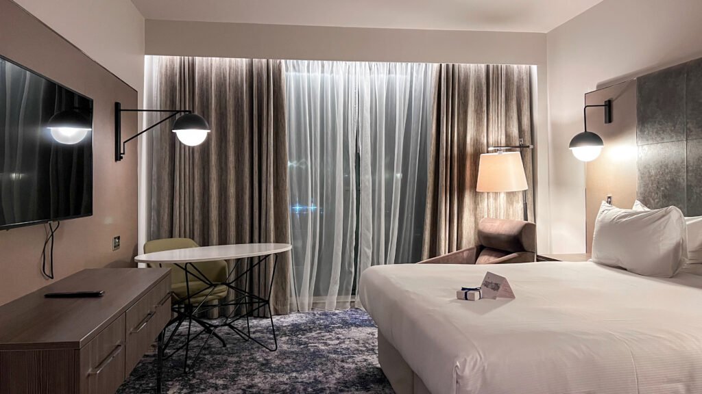 Hilton Aberdeen TECA | Krista the Explorer Hotel room with a king bed, wall lamps, a dresser, and a small round table.
