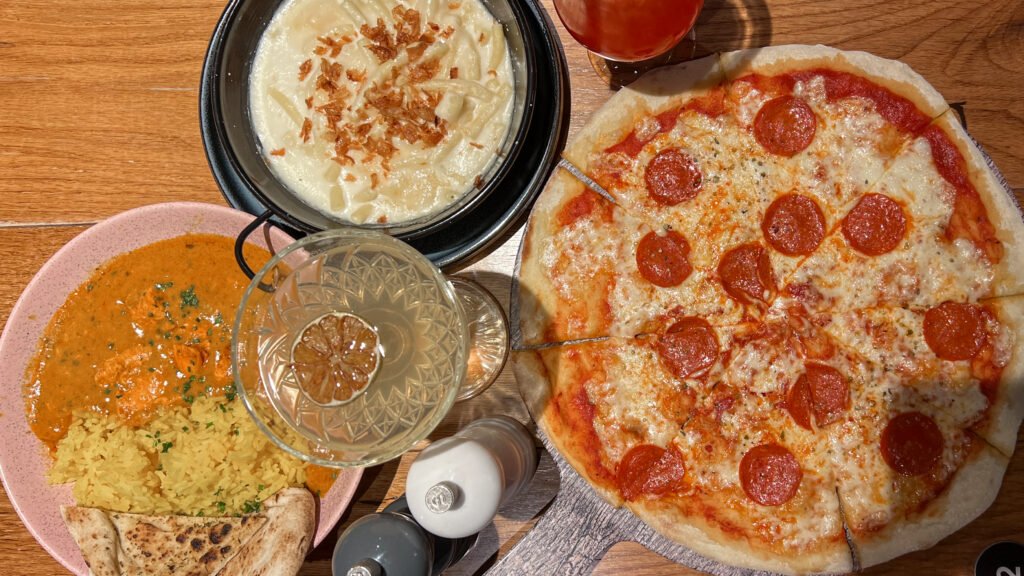 W XYZ Bar Aberdeen | Krista the Explorer Pepperoni pizza on a wooden table with a side of creamy pasta topped with fried onions and a plate of yellow rice with bread.