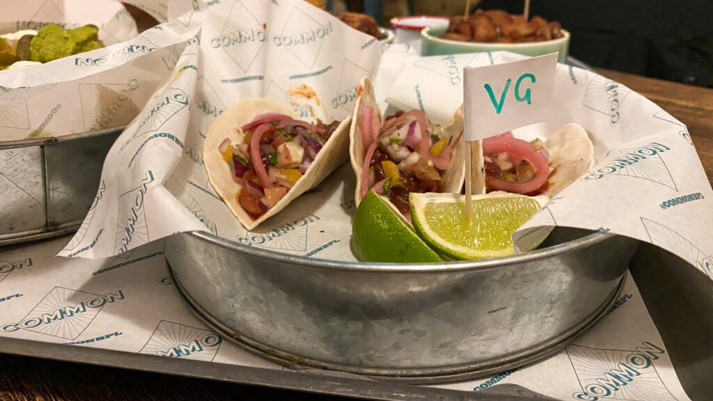 Common Blackpool | Krista the Explorer Two soft-shell tacos filled with diced meat, onions, peppers, and herbs on a metal tray with lime wedges.