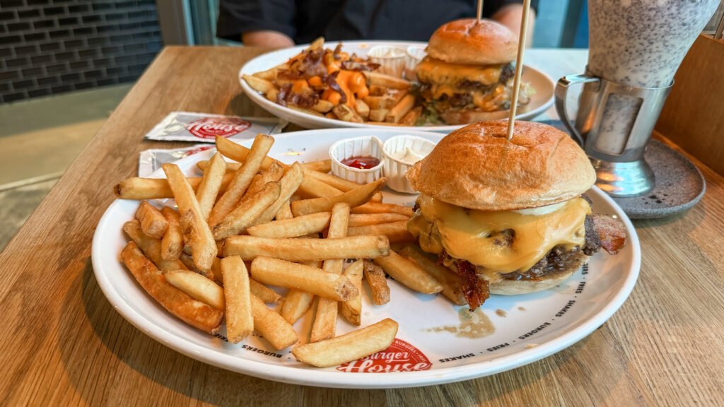 Burger House Trier | Krista the Explorer Cheeseburger with melted cheese and bacon, fries on a plate with condiments nearby on a wooden table.