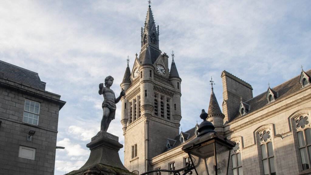 The Mannie Well Aberdeen | Krista the Explorer Clock tower with a tall spire and clock faces, rising behind a stone statue on a pedestal against a blue sky.