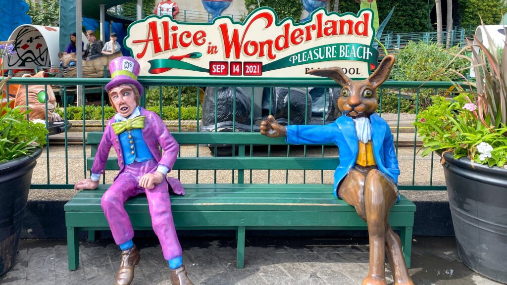 Blackpool Pleasure Beach | Krista the Explorer Statue figures of the Mad Hatter in a purple suit and the White Rabbit in a blue jacket sit on a green bench.