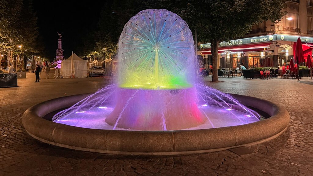 Reims Night | Krista the Explorer Night scene of a rainbow-lit fountain sculpture in a cobblestone plaza.