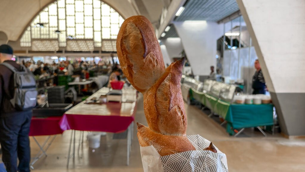 Halles du Boulingrin Reims | Krista the Explorer Close-up of a braided, crusty loaf held in a striped bag at a busy indoor market bakery.