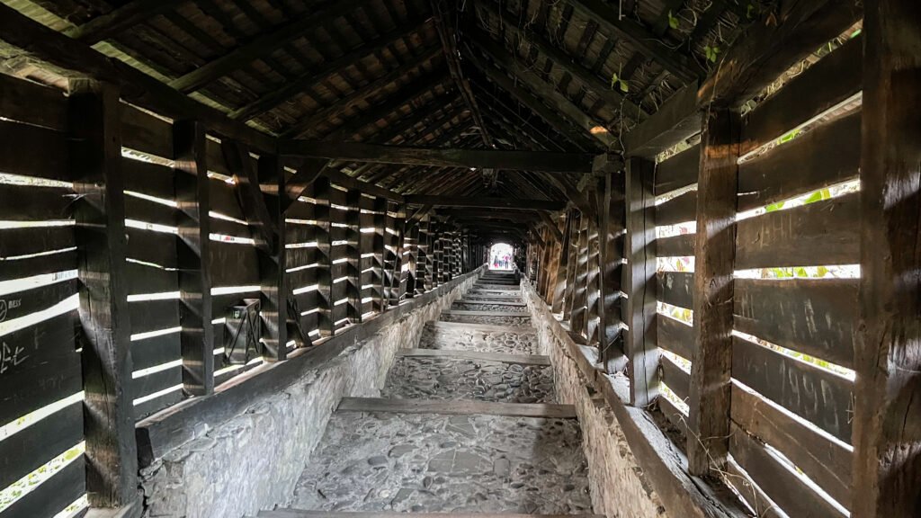 Long, narrow wooden tunnel with slatted walls and a stone floor.
