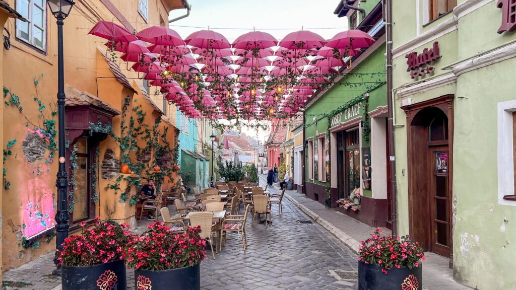Colourful cobblestone street with outdoor cafe seating beneath pink umbrella canopy.
