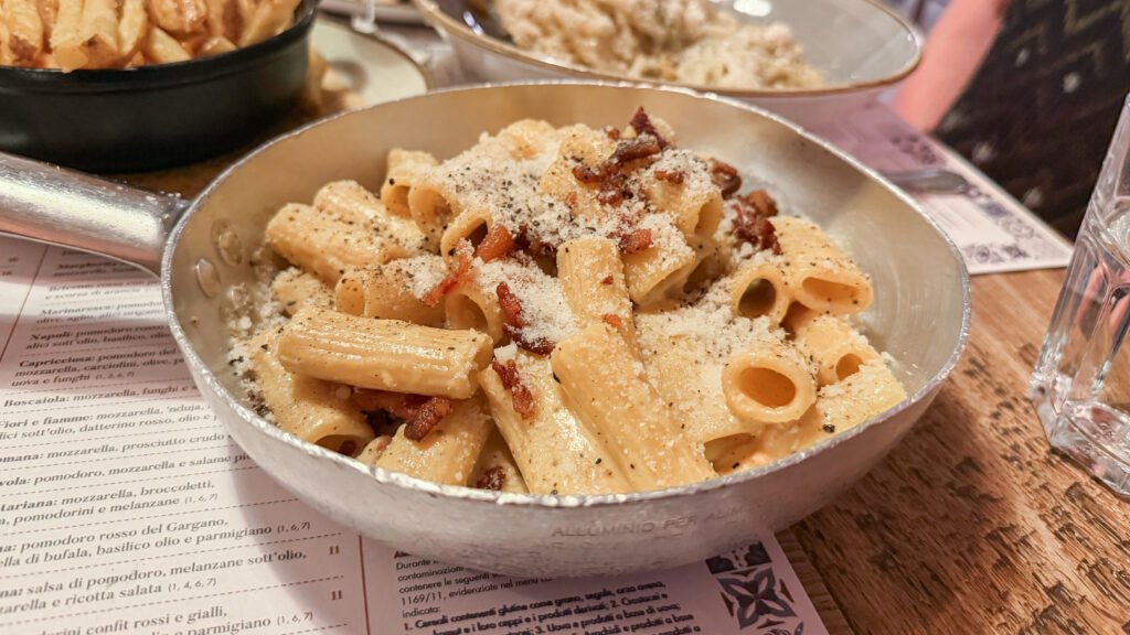 Rione 13 Ristorante Rome | Krista the Explorer Creamy rigatoni pasta with bacon bits and grated cheese in a metal skillet.