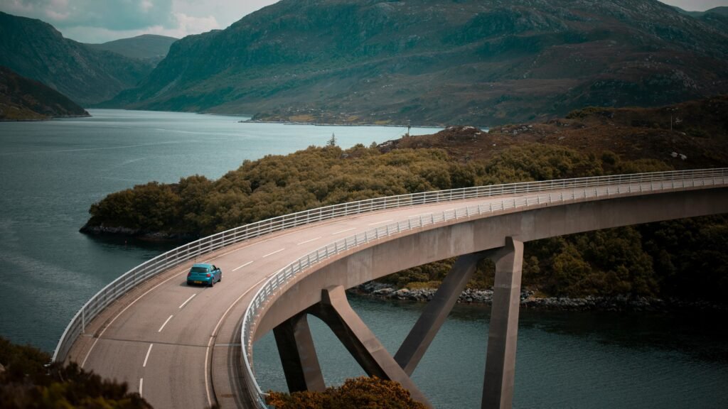 EconomyBookings | Krista the Explorer Curved bridge over a turquoise fjord with green hills and mountains in the distance, a teal car on the road.